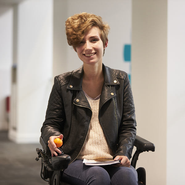 Photo of Jude; young woman who uses  a powered wheelchair with a notebook in her lap.